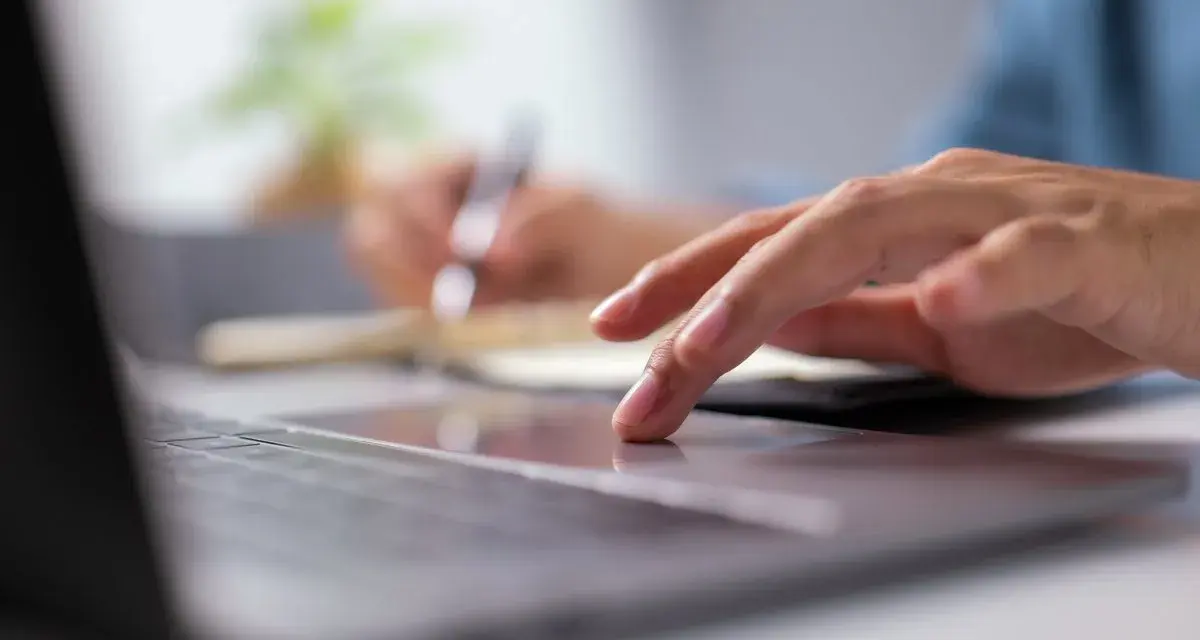 Dłoń na touchpadzie laptopa, w tle notatnik i długopis.