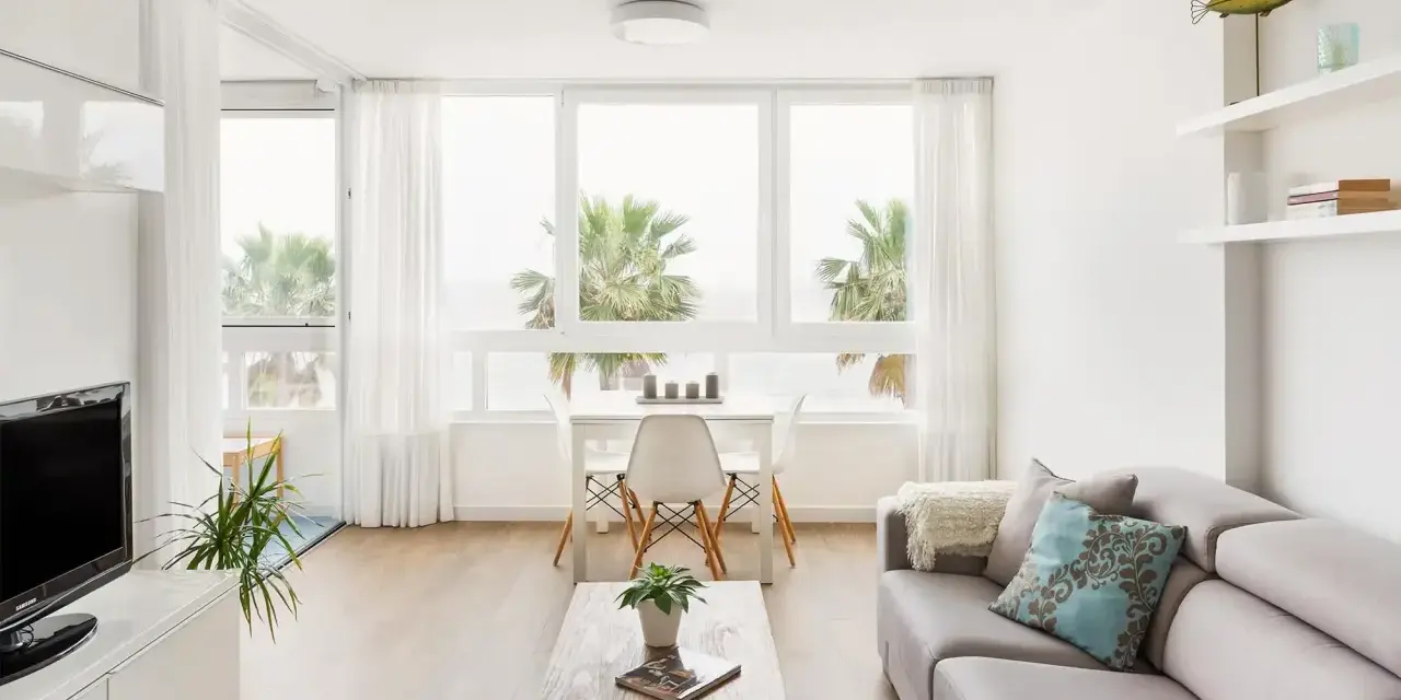 Un luminoso salón estilo nórdico con sofá gris, mesa de centro de madera y grandes ventanales con vistas a palmeras.