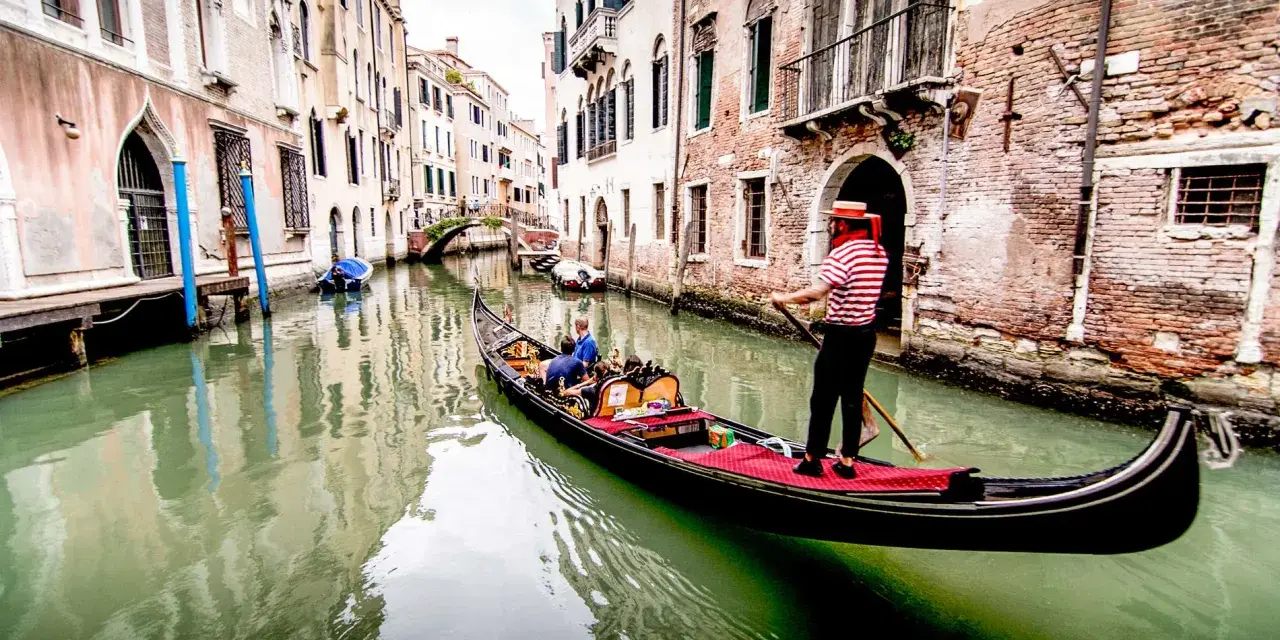Gondoliere in abito a righe guida una gondola con turisti lungo un canale veneziano. Chissà quanto costa un giro in gondola a Venezia per vivere questa magia.
