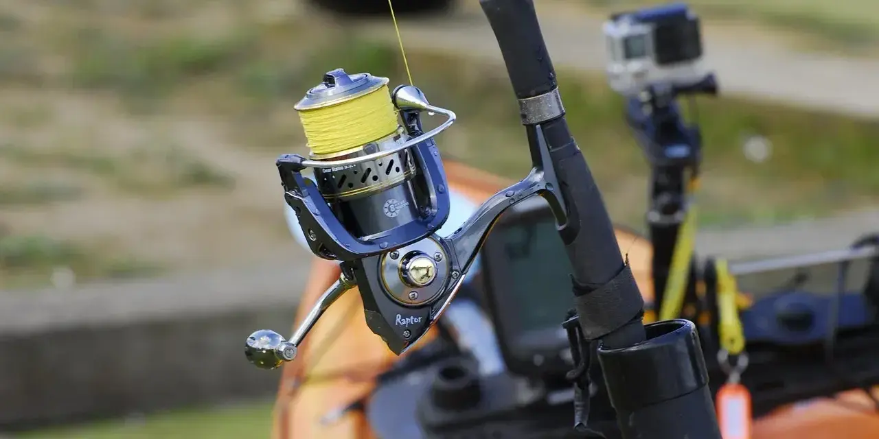 Niebieski kołowrotek spinningowy z żółtą żyłką, gotowy do akcji. W tle widać kajak i kamerę GoPro.