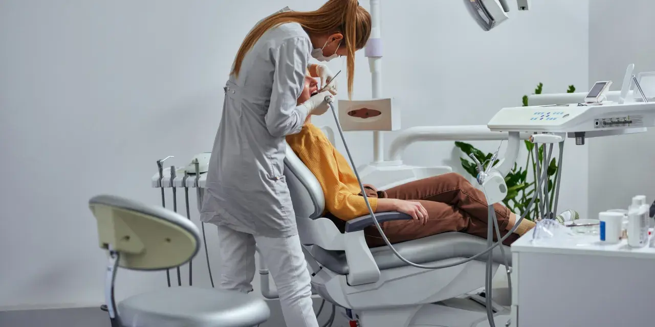 Dentystka w masce i rękawiczkach wykonuje zabieg pacjentowi siedzącemu na fotelu dentystycznym.
