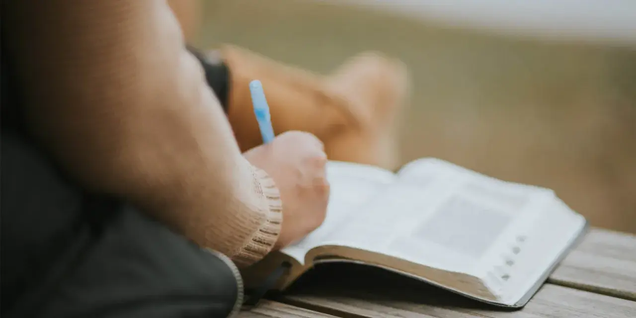 Osoba pisze w otwartej księdze, być może dziennik. W tle widać fragmenty ubrania. To moment refleksji, gdzie żal doskonały może znaleźć swoje ujście w słowach.