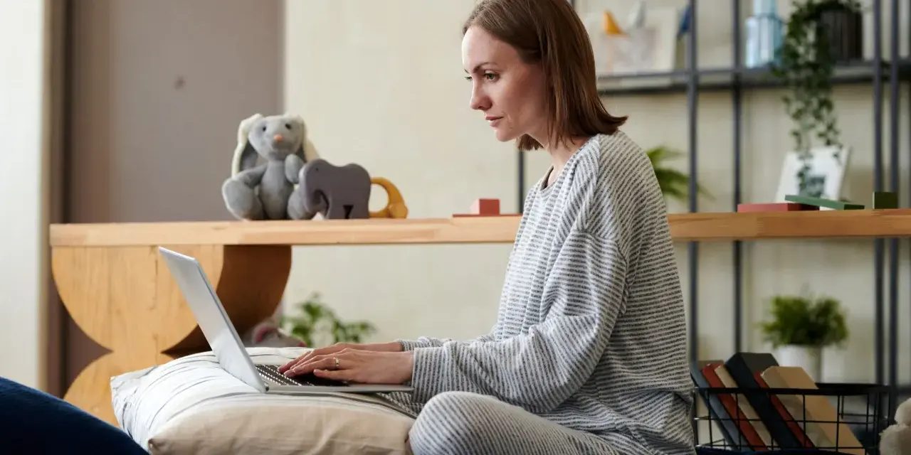 Kobieta w piżamie pisze na laptopie, przygotowując wniosek o urlop macierzyński. Obok pluszowy królik i zabawki.