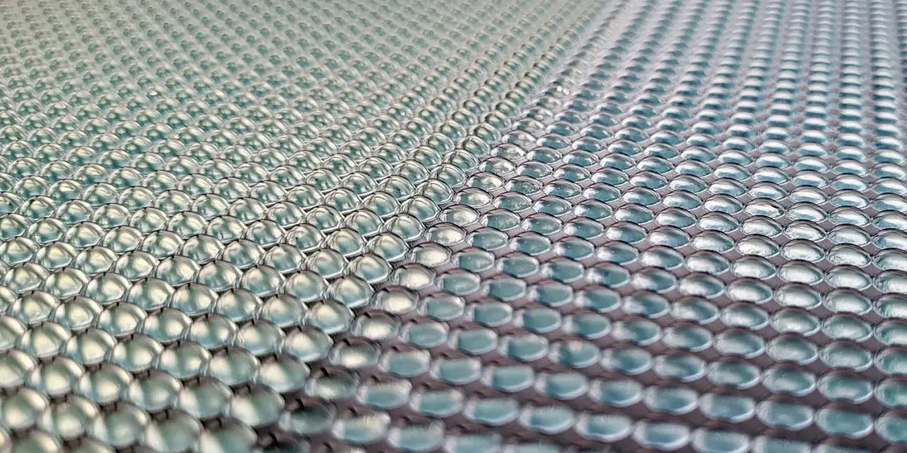 Close-up of a 3d printed lens array, with rows of tiny, clear, dome-shaped lenses reflecting light.