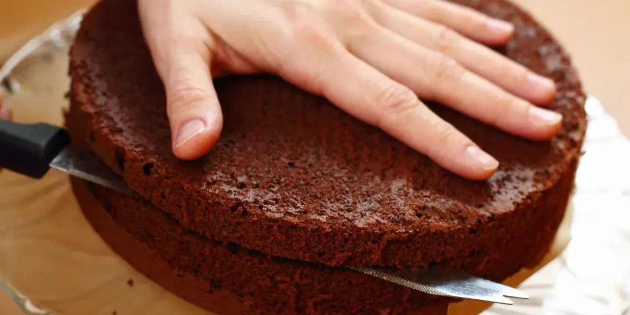 Eine Hand hält einen Schokoladenkuchen, während ein Messer ihn waagerecht durchschneidet, um den tortenboden zu teilen.