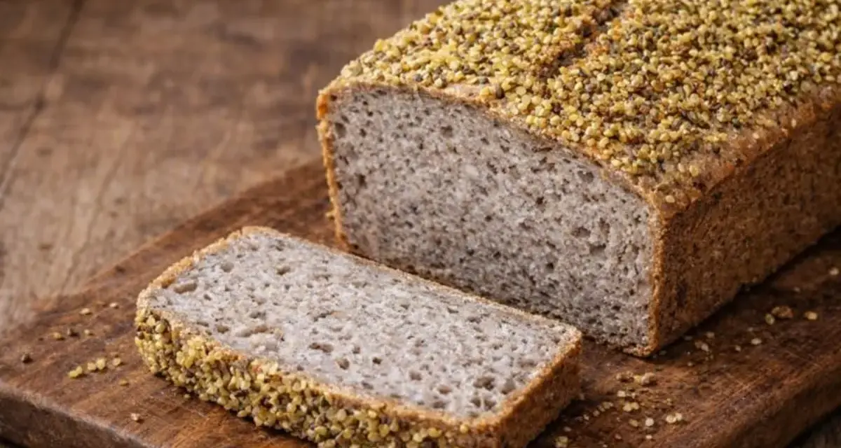 Ein rustikales Buchweizenbrot mit einer Scheibe auf einem Holzbrett. Die Kruste ist mit Buchweizenkörnern bestreut.
