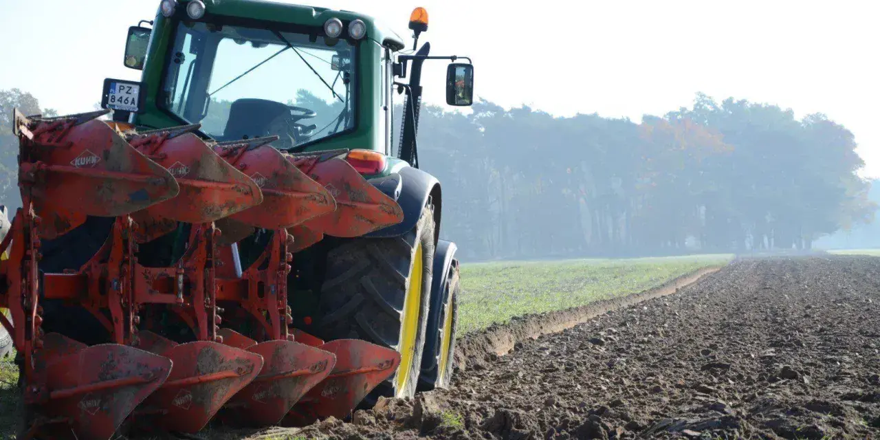 Zielony traktor z pługiem orze pole. W tle mglisty las.