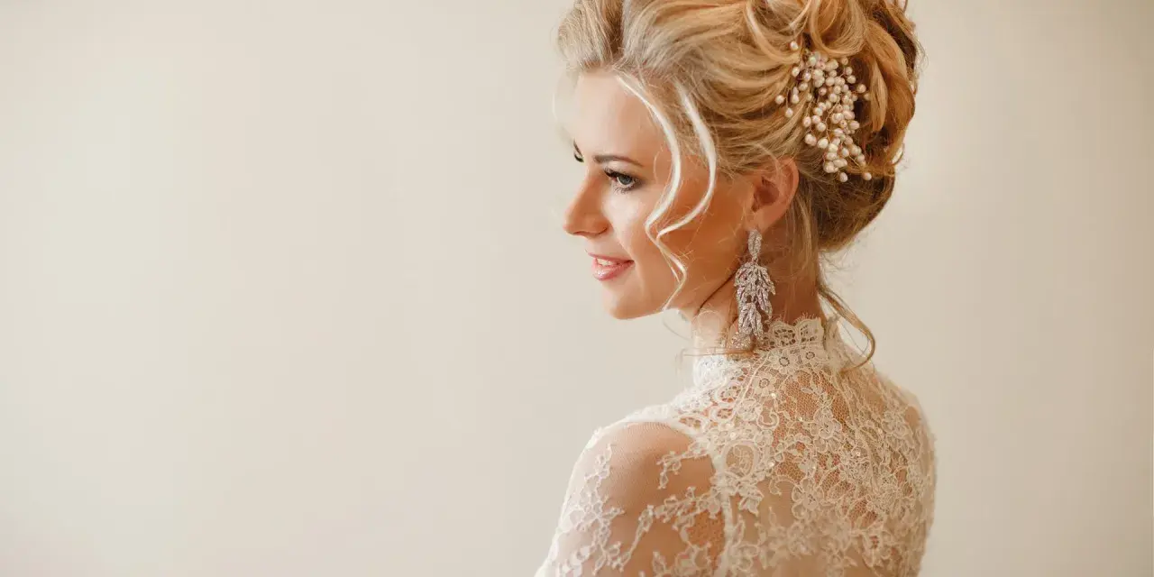 Eleganckie uczesania na wesele średnie włosy: blondynka w koronkowej sukni z perłową ozdobą i długimi kolczykami.