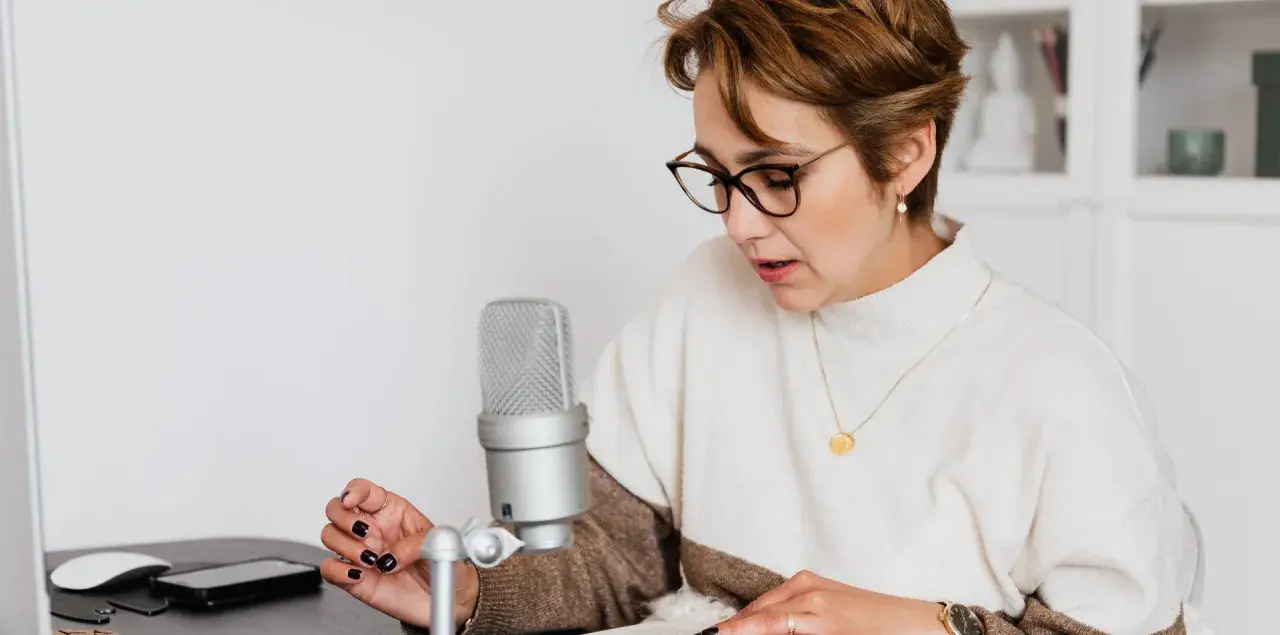Mujer con gafas lee un libro frente a un micrófono, aprendiendo como escribir mejor.