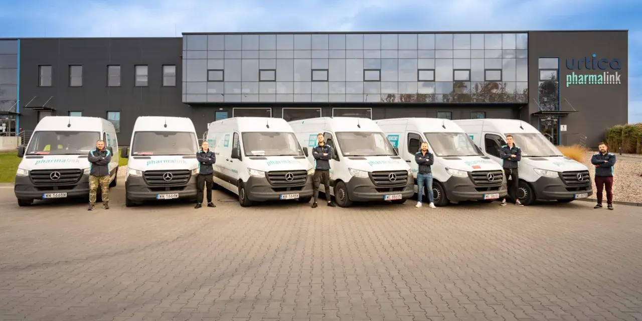 Sześciu mężczyzn w uniformach stoi przed rzędem białych vanów z logo "pharmalink". To zespół odpowiedzialny za transport leków, profesjonalny przewoźnik.