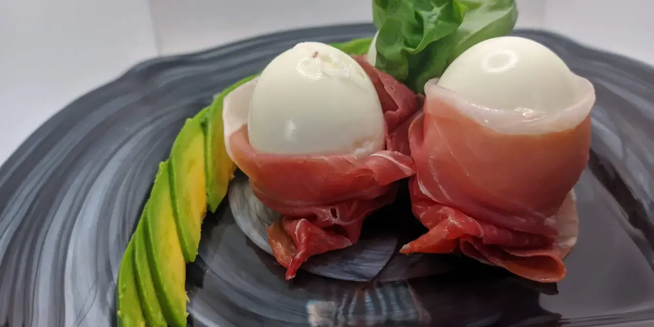 Due uova di quaglia sode avvolte in prosciutto crudo, servite con fette di avocado e foglie verdi su un piatto scuro.