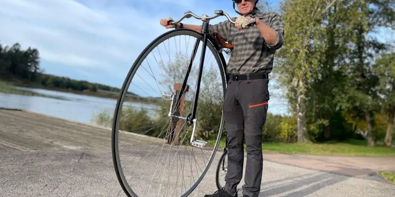 Man med hjälm och solglasögon står bredvid en cykel med stort framhjul vid en sjö.