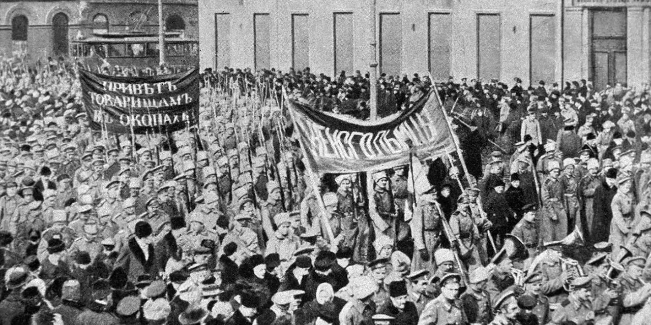 Multitud con pancartas y uniformes militares marchando en una calle, con edificios al fondo.