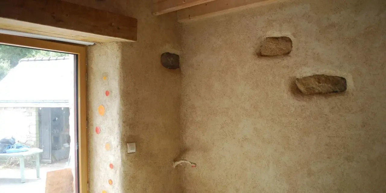 Un mur avec un enduit chaux finition texturé, intégrant des pierres apparentes et des touches de couleur. Le bois des poutres et du cadre de fenêtre apporte de la chaleur.