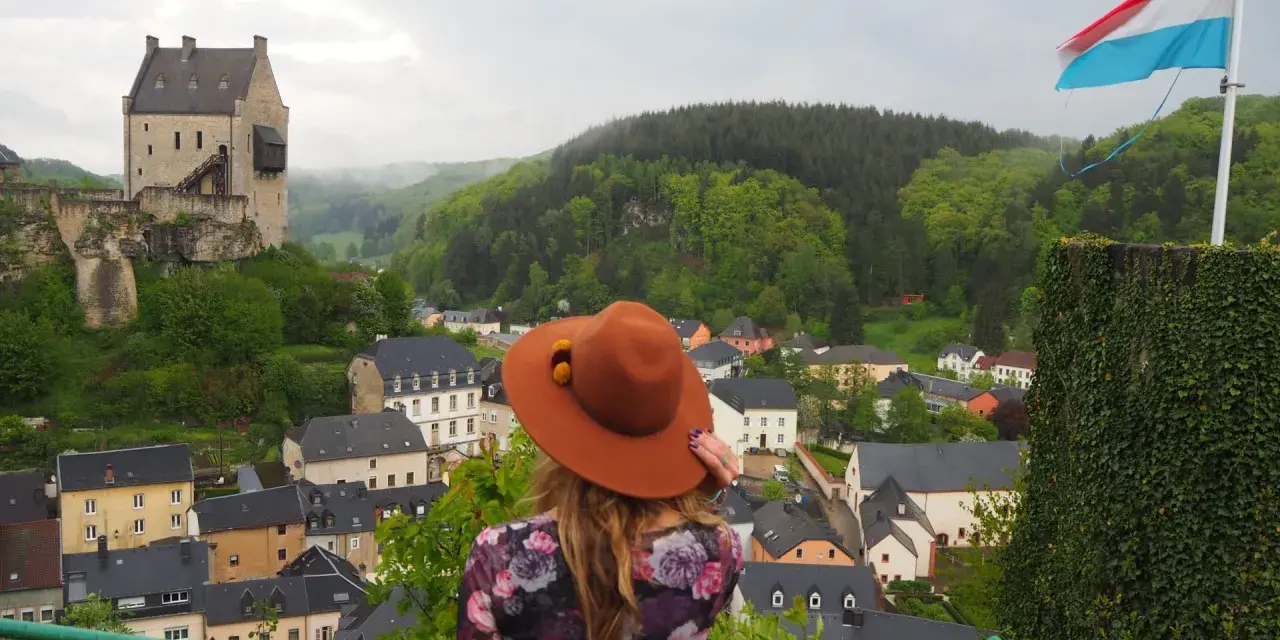 Mujer con sombrero marrón admira un castillo y un pueblo, con la bandera de Luxemburgo ondeando. ¡Un paisaje digno de un buen queso Muenster!