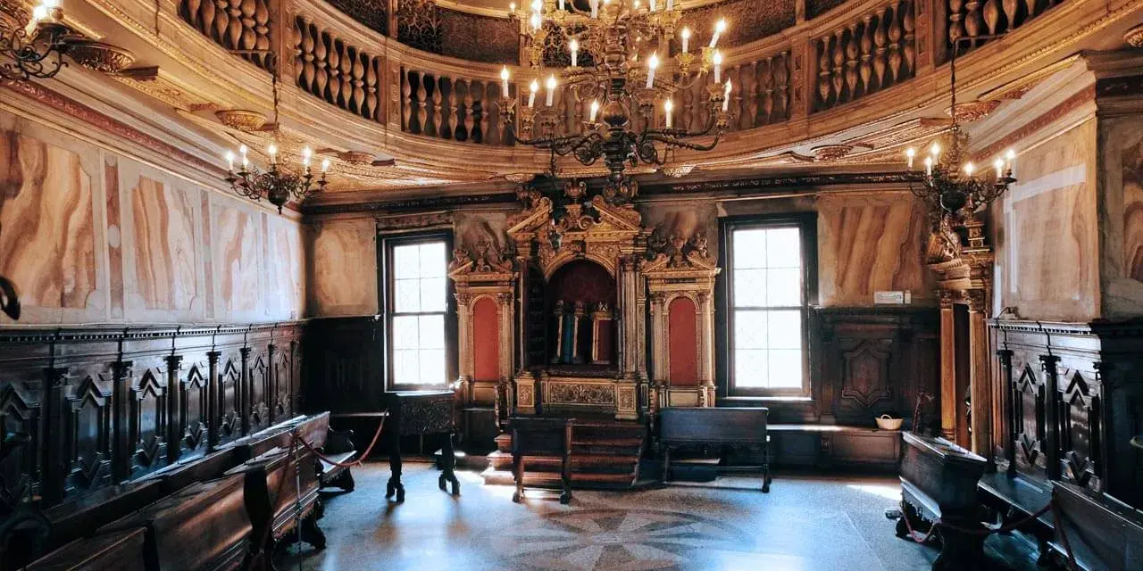 Interno sontuoso della sinagoga di Venezia, con arredi in legno scuro, un grande lampadario e l'Arca Santa dorata.