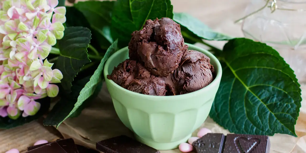Ein grüner Napf mit zartbitter schokoladeneis, umgeben von Schokoladentafeln, rosa Bonbons und einer Hortensienblüte.