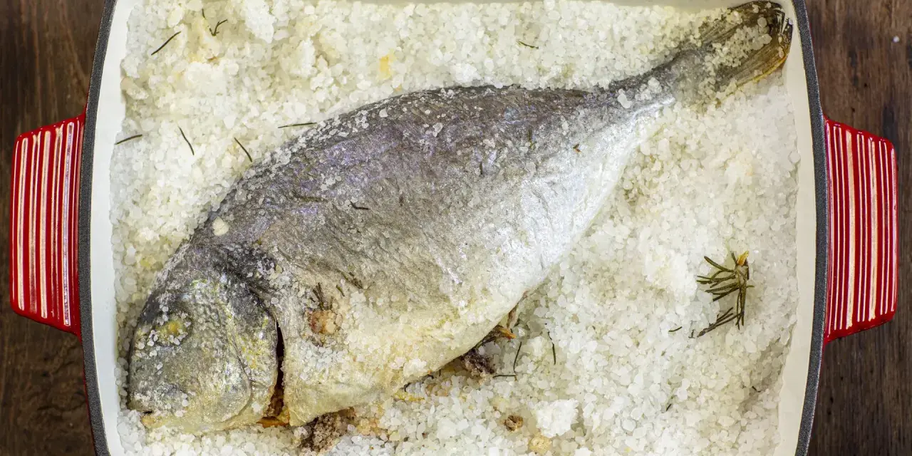 Orata al sale, intera e dorata, adagiata su un letto di sale grosso e rosmarino in una teglia rossa.