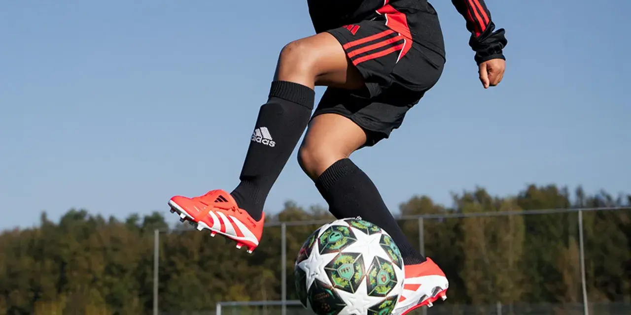 Barn i luften med fotboll. Svarta strumpor med Adidas-logga och benskydd fotboll barn storlek. Orange fotbollsskor med vita detaljer.