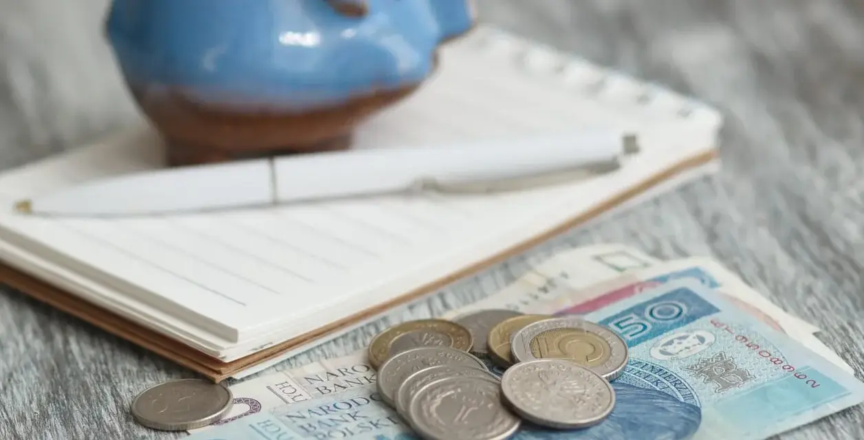 Świnka skarbonka, notatnik i długopis nad polskimi banknotami i monetami. Zastanawiasz się, ile wyniesie waloryzacja kapitału początkowego w 2025?