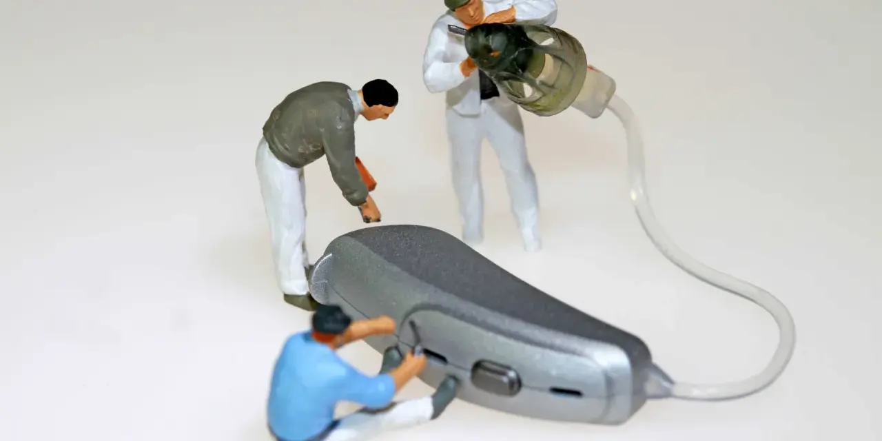 Miniaturowi pracownicy zajmują się naprawą aparatów słuchowych. Jeden z nich przykręca element, drugi czyści słuchawkę, a trzeci pracuje nad aparatem.