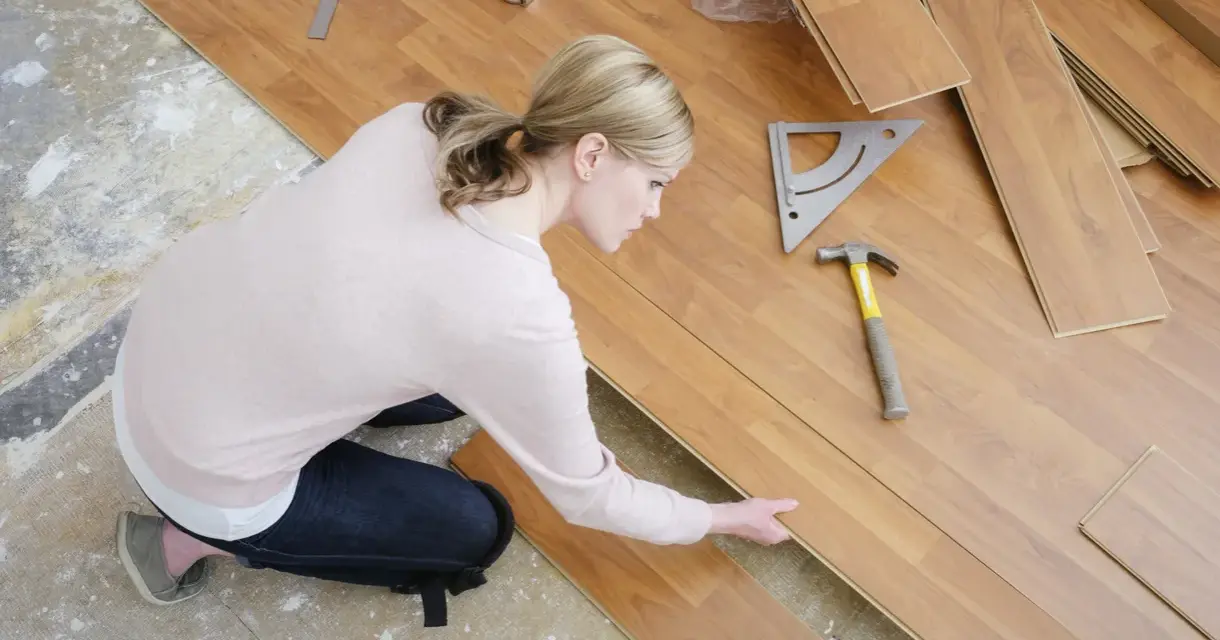 Frau kniet und verlegt Laminat. Werkzeuge wie Hammer und Winkel liegen bereit.