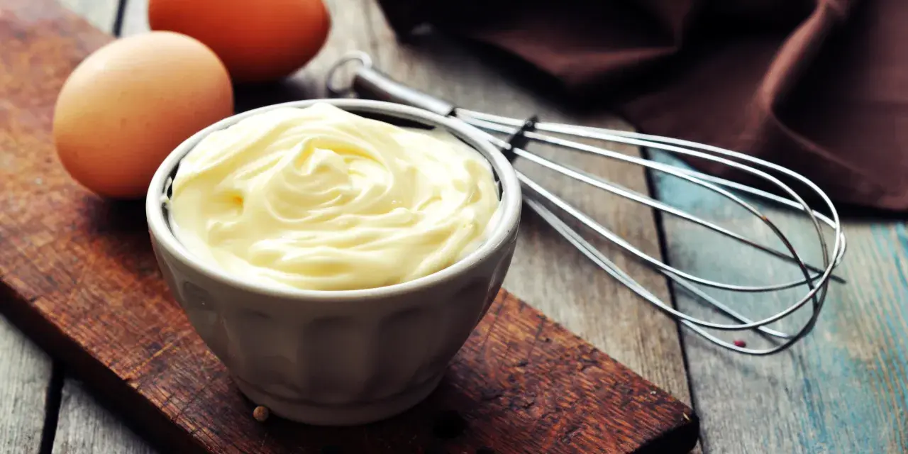 Un bol de mayonnaise onctueuse, des œufs et un fouet, prêts à faire une mayonnaise maison.
