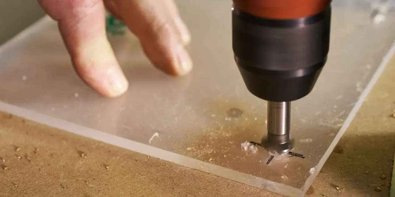 A drill bit is used to drill through acrylic. A hand guides the acrylic sheet as the drill creates a hole, showing it's possible to drill through acrylic.