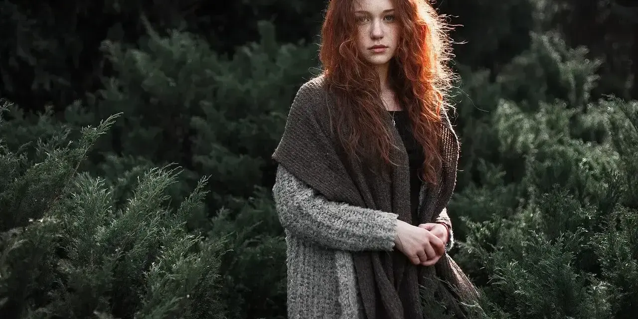 Rudowłosa dziewczyna w grubym swetrze i szalu, otoczona ciemną zielenią. Jej spojrzenie jest spokojne, a cała scena ma **low-key** klimat.