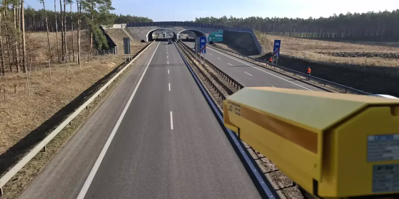 Żółta kamera monitoruje odcinek pomiarowy prędkości na S8. Droga prowadzi przez las, pod wiaduktem.