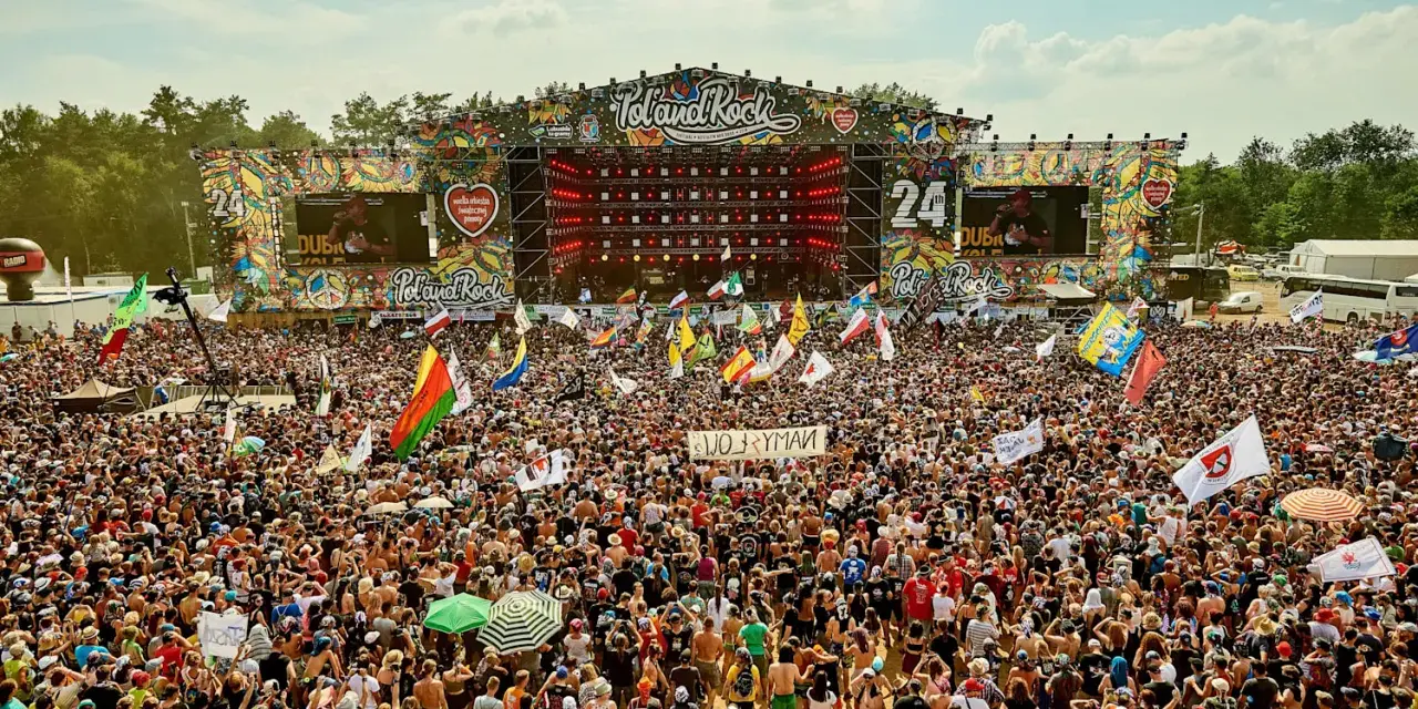Tłum ludzi na festiwalu Poland Rock, gdzie jest scena i ekrany. Wszędzie flagi i słońce.