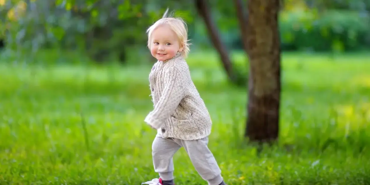 Ein fröhliches Kind, ca. 2,5 Jahre alt, mit blonden Haaren und einem Strickcardigan, läuft auf einem Weg im Park.