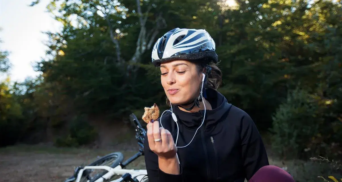 Kobieta w kasku i słuchawkach je ciastko, obok leży rower. To przykład zdrowego odżywiania i aktywnego stylu życia, czyli zbilansowana dieta.