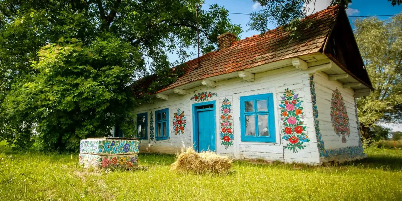 Malowany dom w Zalipiu, z niebieskimi oknami i drzwiami, ozdobiony kwiatami. Obok studnia i snop siana. Cennik wizyty w tym malowniczym miejscu jest dostępny dla zwiedzających.