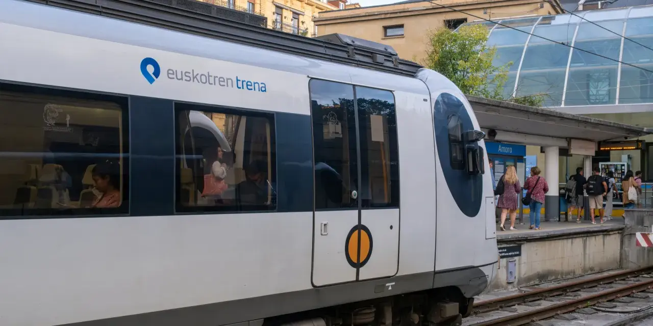 A modern Euskotren train at San Sebastian train station. Passengers wait on the platform under a glass-covered canopy.