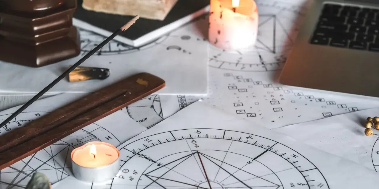 Na stole leżą wykresy astrologiczne, świece, kadzidełko i laptop. Widać symbolikę wskazującą na ascendent w bliźniętach.