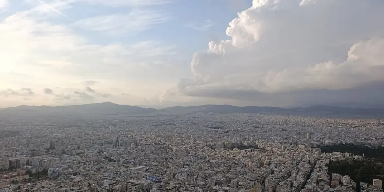 Rozległa panorama Aten z wzgórza Lycabettus. Miasto rozciąga się aż po horyzont, a nad nim piętrzą się chmury.