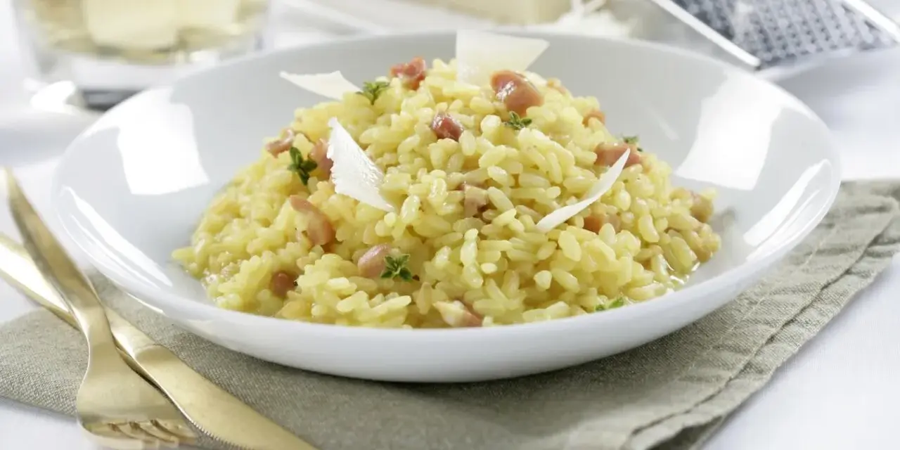 Un cremoso arroz a la milanesa, adornado con láminas de queso parmesano y hierbas frescas, servido en un plato blanco.