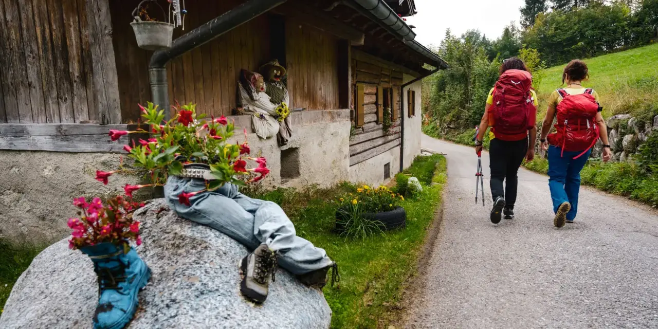 Due escursioniste affrontano il cammino San Vili, con zaini rossi e bastoncini. La strada, tra case rurali e verde, suggerisce una certa difficoltà.