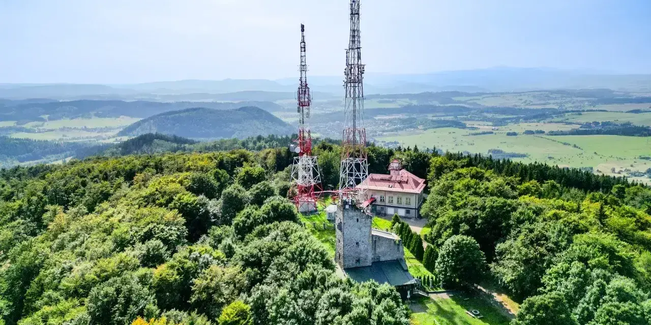 Wieże telekomunikacyjne na zalesionym szczycie Chełmiec. W tle rozciąga się malowniczy krajobraz pogórza.