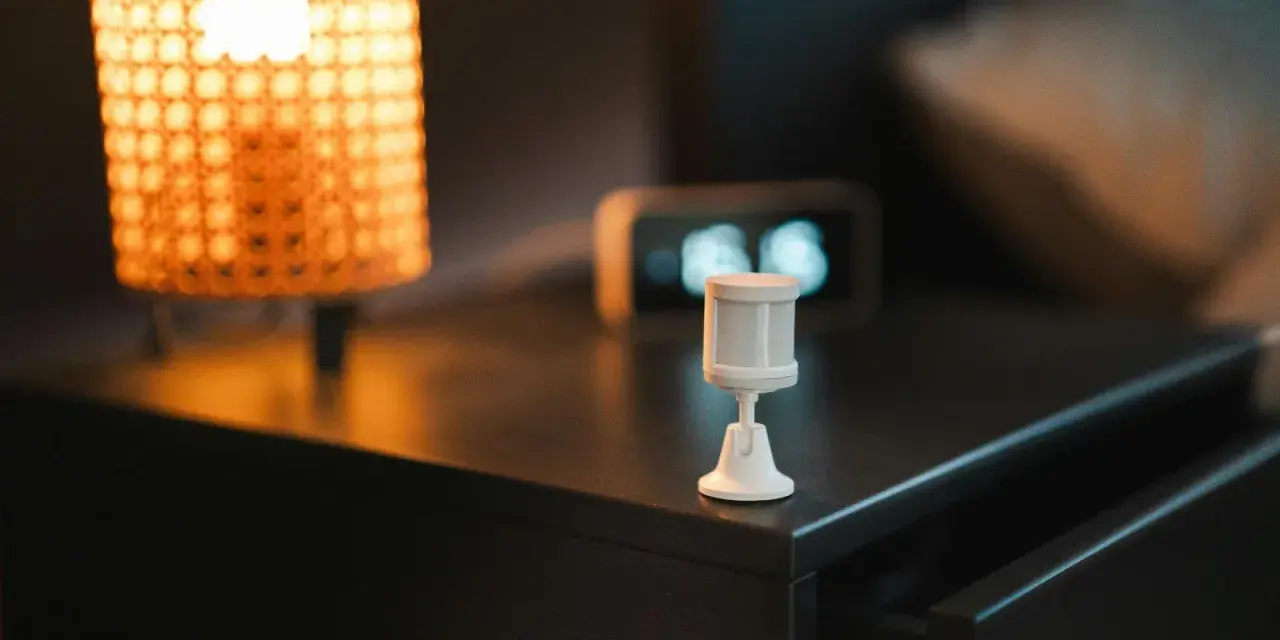 Sensor de movimiento blanco sobre mesita de noche, junto a lámpara y reloj. Ideal para saber como dejar fijo un sensor de movimiento.