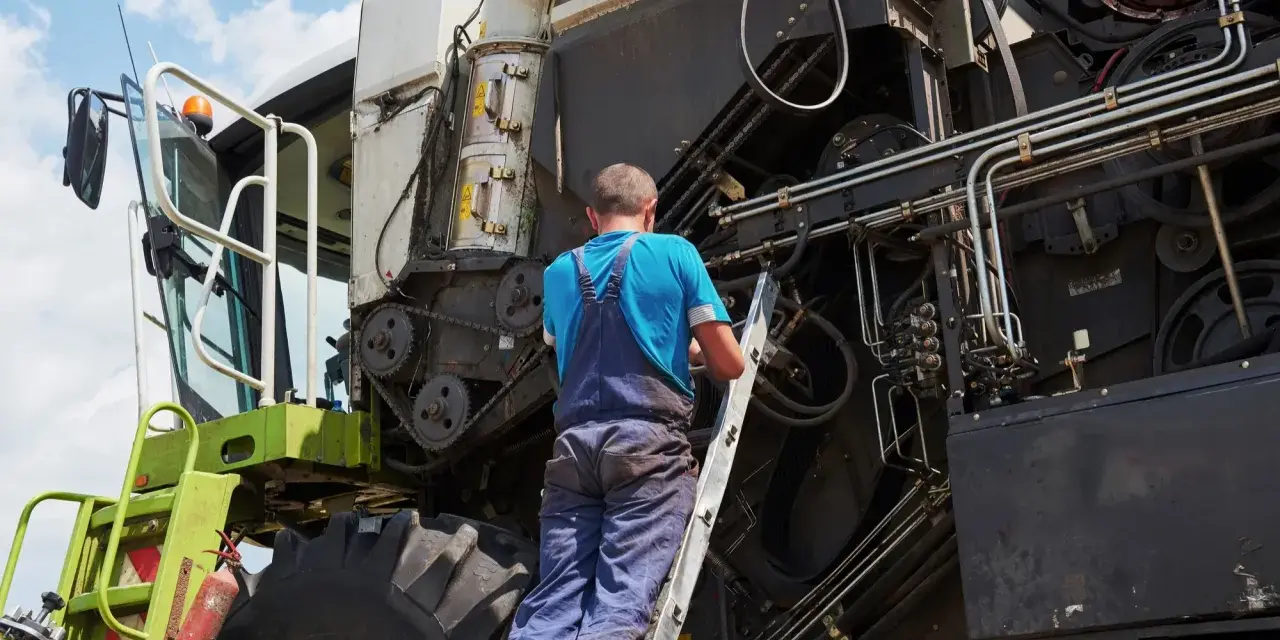 Mechanik na drabinie naprawia kombajn. Co to jest tuz? To potężna maszyna rolnicza, która zbiera plony.