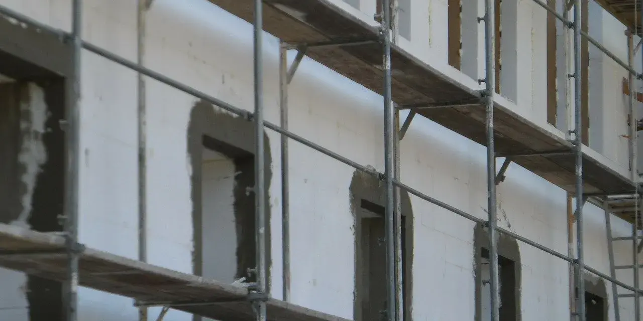 Budowa domu z bloczków styropianowych. Rusztowania otaczają białą ścianę z otworami okiennymi i drzwiami, czekającymi na wykończenie.