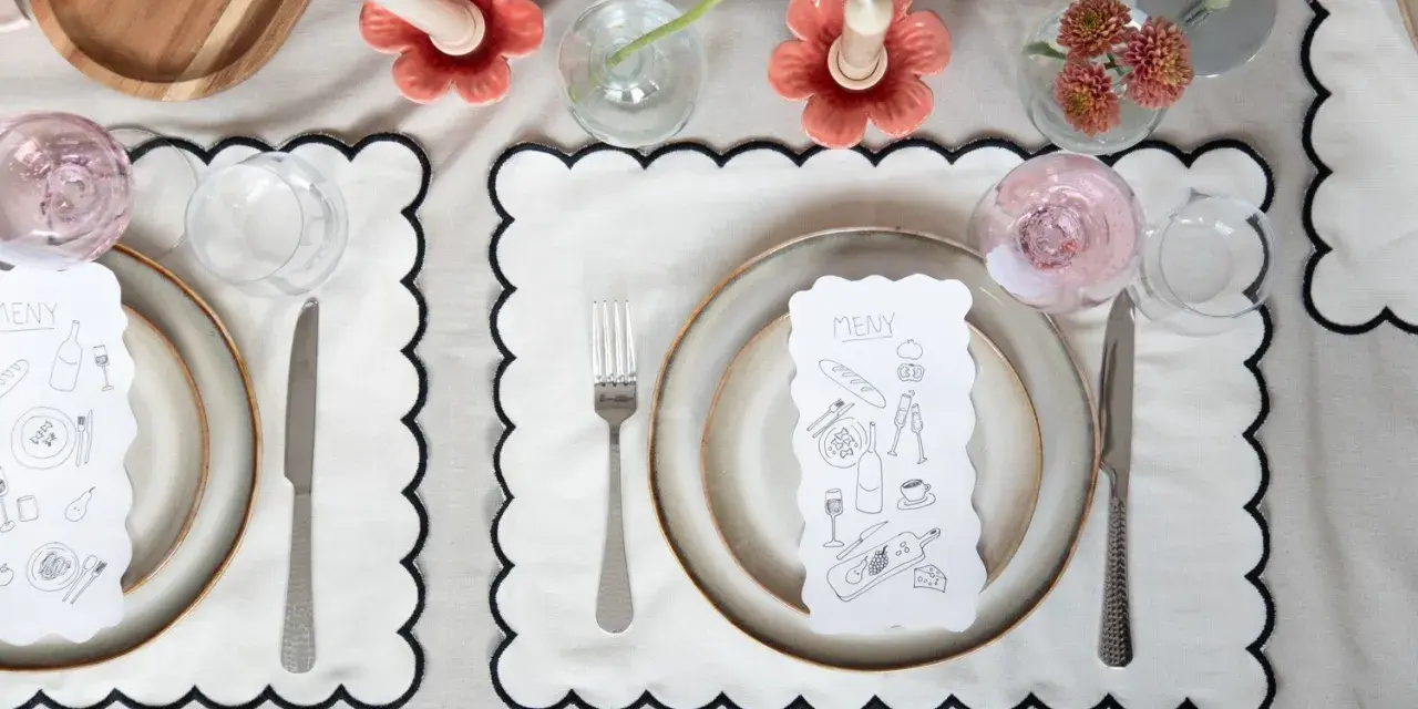 A table setting with a gold-colored tablecloth, white placemats with black scalloped edges, plates, cutlery, glasses, and a menu with illustrations.