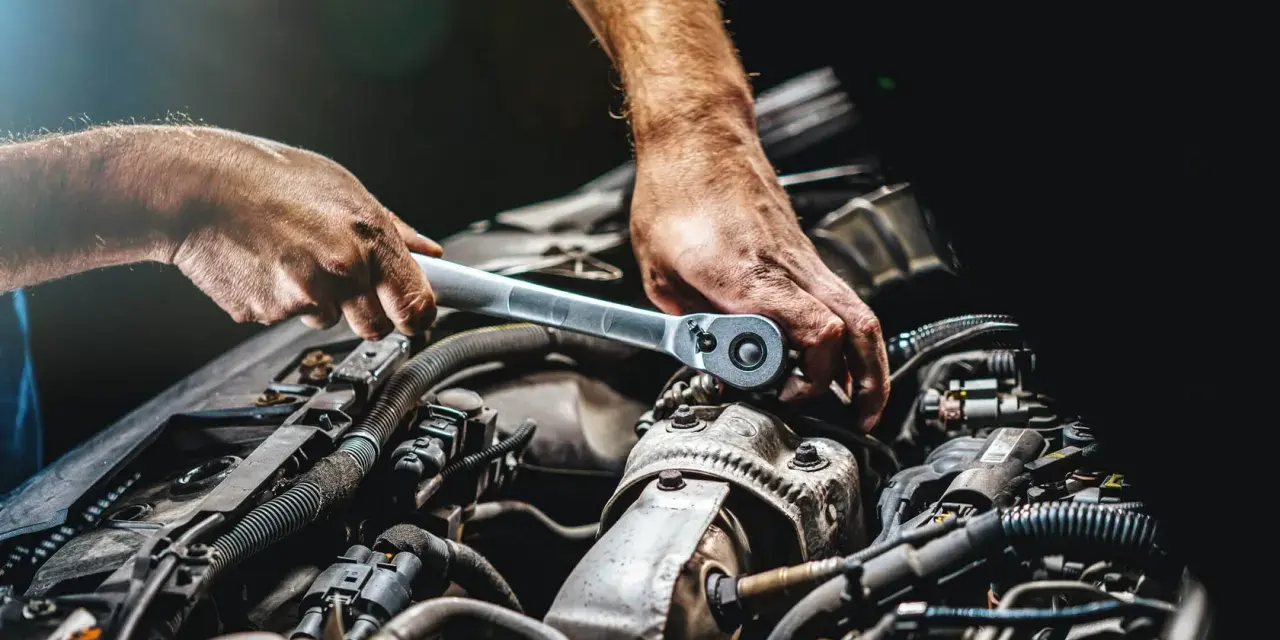 Mechanik dokręca śrubę kluczem w silniku samochodu. Widać jego dłonie i fragmenty silnika, być może podczas rutynowej kontroli lub naprawy.