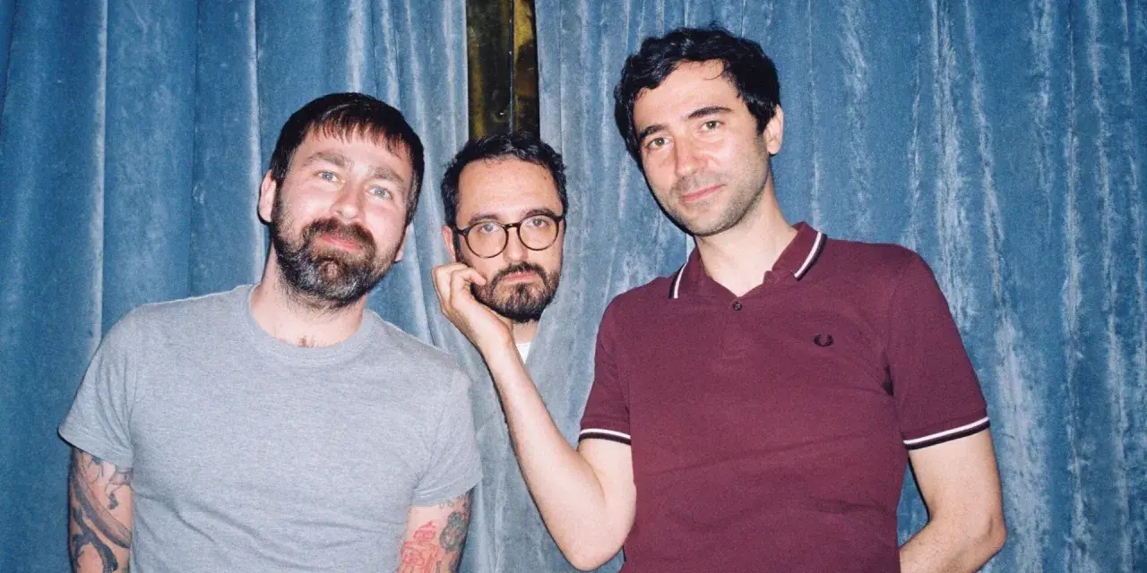 Trois hommes posent devant un rideau bleu. L'un d'eux, au centre, est un groupe musique espagnol année 2000.