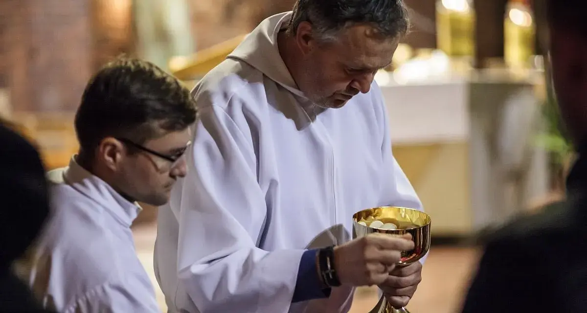 Szafarz, w białej albie, trzyma złoty kielich z hostiami. Obok niego młodszy mężczyzna w okularach.