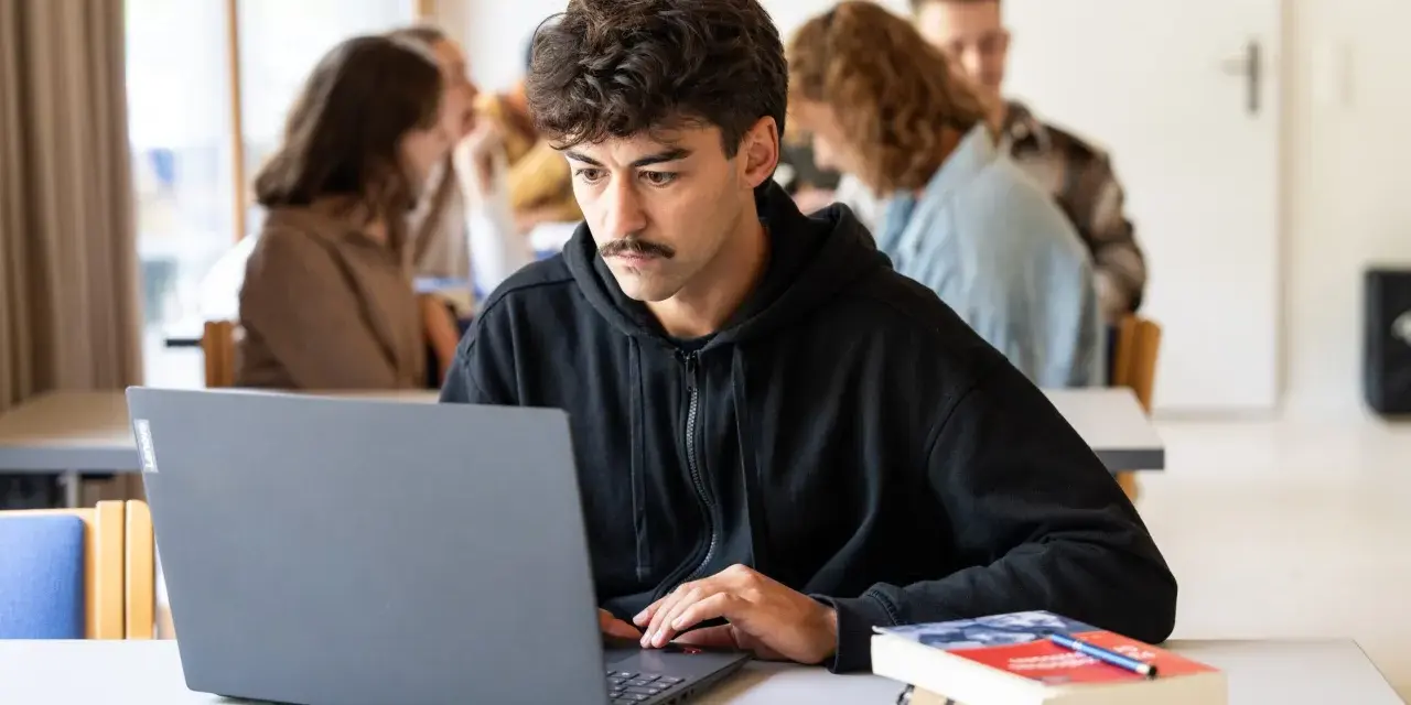 Ein junger Mann mit Schnurrbart konzentriert sich auf seinen Laptop, während er ein evangelisches Fernstudium Theologie absolviert.