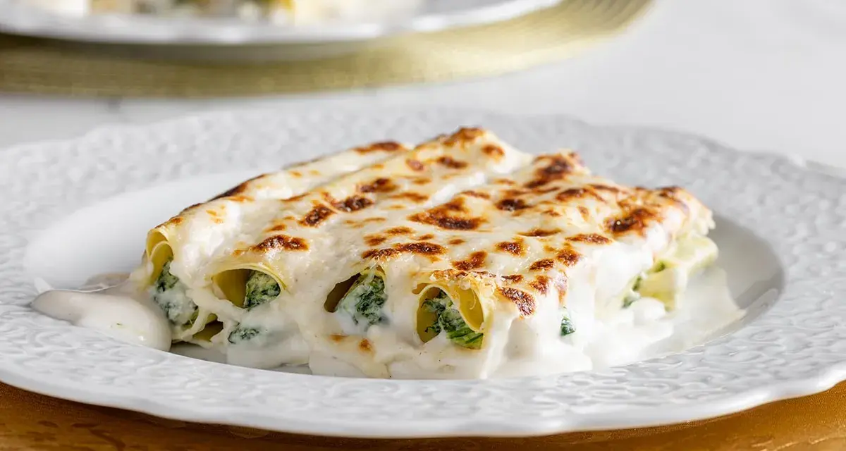 Cannelloni ricotta e spinaci fumanti, ricoperti da besciamella e formaggio fuso, serviti su un piatto bianco.
