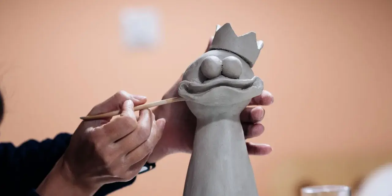 Manos trabajando con arcilla para crear una figura de rana con corona.